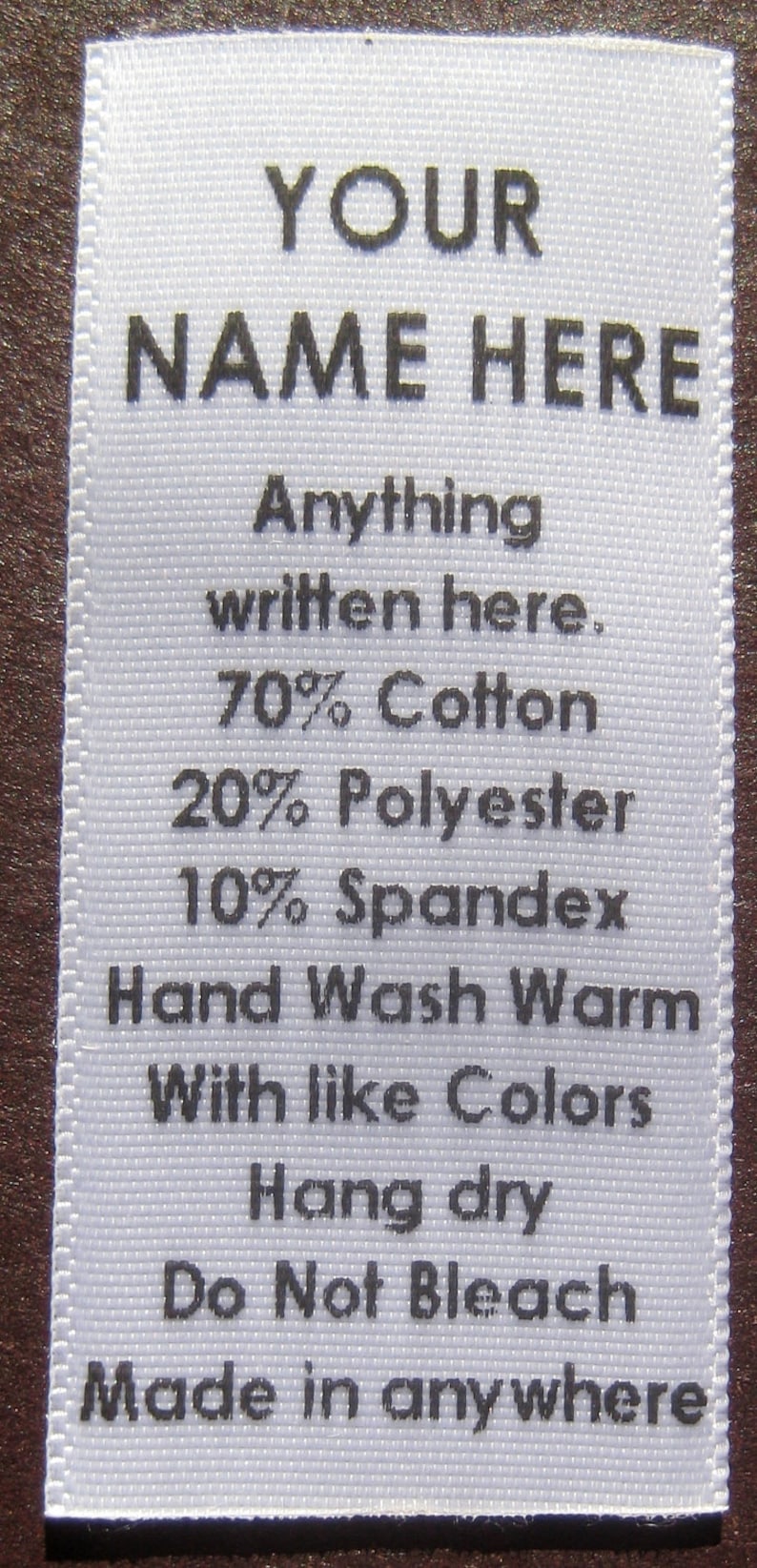 May include: White clothing label with black text. The text reads "YOUR NAME HERE", "Anything written here", "70% Cotton", "20% Polyester", "10% Spandex", "Hand Wash Warm", "With like Colors", "Hang dry", "Do Not Bleach", and "Made in anywhere".