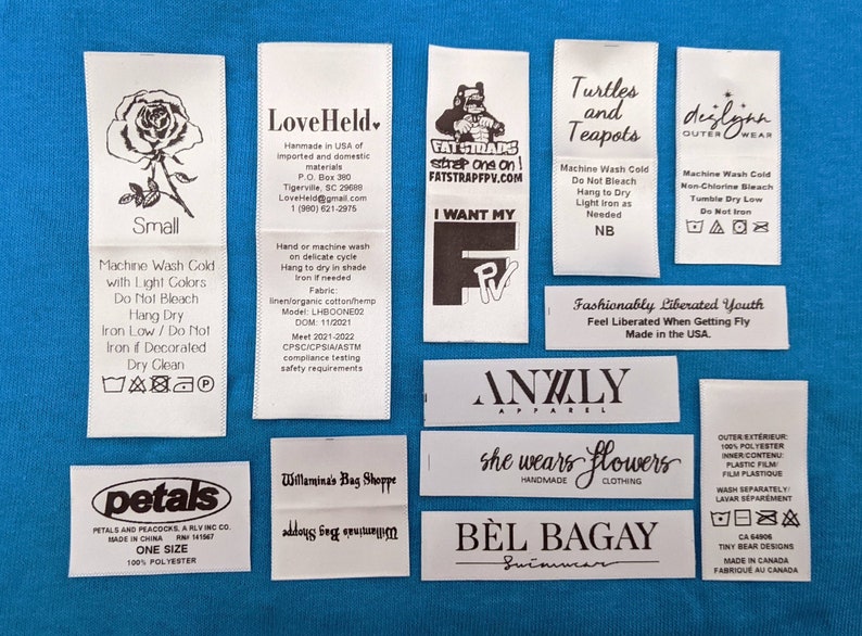 May include: A collection of white fabric clothing labels with black text and graphics. The labels include brand names, care instructions, size information, and fabric content. Some of the labels feature a rose, a turtle, a teapot, and a stylized illustration of a person.