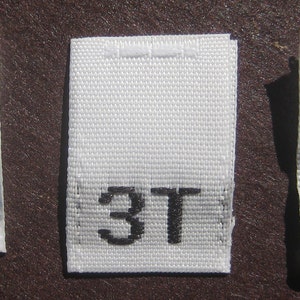 Könnte beinhalten: Drei weiße Stoff-Etiketten mit schwarzem Text. Die Etiketten sind mit "2T", "3T" und "4T" beschriftet.
