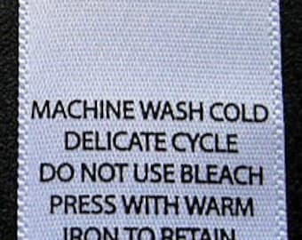 Op de afbeelding: Wit waslabel met zwarte tekst met instructies voor het wassen en strijken van een kledingstuk. De instructies luiden: Machinewas koud, delicate cyclus, niet bleken, strijken met warm strijkijzer om de vorm te behouden.