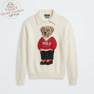 May include: A cream-colored long-sleeve knit sweater with a collared neckline. The center features an intarsia-knit teddy bear wearing a red hooded sweatshirt with the word POLO in white, black pants, and red and black sneakers.