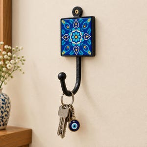 May include: A black metal wall hook featuring a square decorative tile with a blue, yellow, and white floral mandala pattern. The hook holds a silver metal keyring with two keys and a small circular charm with a blue and white evil eye design.