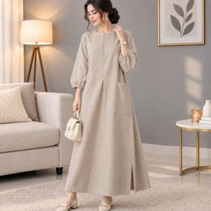 May include: A long-sleeved, floor-length beige dress featuring a round neckline, a single large patch pocket on the front, and a side slit at the hem. The dress has a straight silhouette with gathered cuffs at the wrists and a subtle center seam detail.