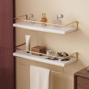 Puede incluir: Dos estantes de pared rectangulares blancos con barandillas de metal dorado. El estante superior sostiene tres frascos de perfume de vidrio. El estante inferior sostiene un tubo, una caja de madera, un frasco pequeño, un accesorio de tela y una toalla blanca.