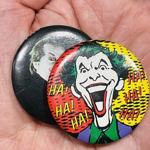 May include: Two circular pin-back buttons featuring The Joker. One button displays a black and white photographic portrait of a man. The other button features a colourful comic-style illustration of The Joker with green hair and the text HA! HA! HA!