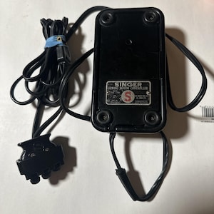 Peut inclure: Un contrôleur de moteur de machine à coudre Singer noir avec une base rectangulaire, quatre pieds circulaires et un cordon électrique avec une prise à trois broches. Une plaque métallique porte le texte SINGER SEWING MOTOR CONTROLLER et SIMANCO.