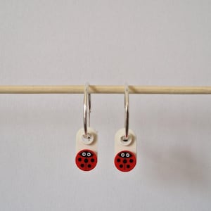 Op de afbeelding: Een paar zilveren oorringen met kleine, gebroken witte rechthoekige bedels. Elke bedel is versierd met een cirkelvormig rood lieveheersbeestjesmotief met zwarte stippen en twee witte ogen, hangend aan de dunne zilveren metalen ring.