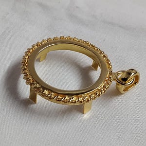 May include: A gold-toned circular pendant setting featuring a twisted rope-style border, a smooth inner rim, and four vertical prongs extending downward to secure a gemstone. A small, polished gold-toned bail is attached to the side.