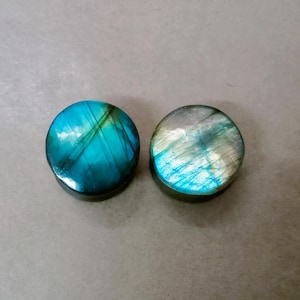Può includere: Due dilatatori circolari con facce in pietra labradorite lucida. Il dilatatore a sinistra mostra un'iridescenza blu ottanio profonda con inclusioni scure, mentre quello a destra presenta un riflesso argentato e blu più chiaro e levigato.