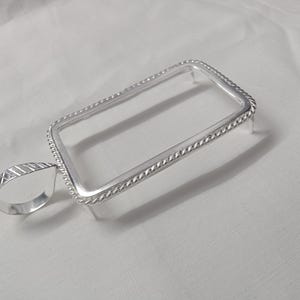 May include: A rectangular silver pendant setting featuring a twisted rope-style border and a smooth, teardrop-shaped bail with diagonal etched lines. The frame includes four small prongs extending downward from the corners for securing a stone.