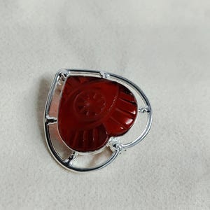 Può includere: Una spilla a forma di cuore con un centro in pietra intagliata rosso scuro traslucido, incastonata in una cornice di metallo color argento lucido. La pietra presenta un motivo radiale a raggiera inciso sulla superficie, circondato da un bordo liscio.