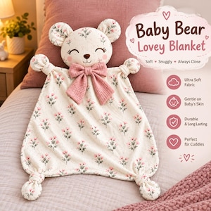 May include: A cream-coloured baby bear lovey blanket featuring a pink floral pattern, a bear head with closed eyes, and a large pink bow at the neck. The corners are knotted, and the text reads Baby Bear Lovey Blanket, Soft, Snuggly, Always Close.