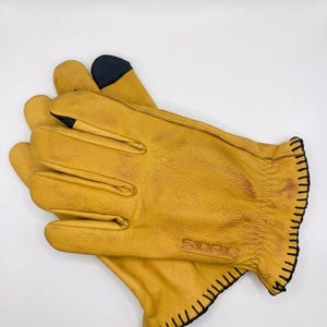 Puede incluir: Un par de guantes de cuero color amarillo mostaza con costuras negras en los puños y detalles en azul marino oscuro en las puntas de los dedos. La palabra SIDRIO está grabada en el dorso de la mano y tienen un acabado natural texturizado.
