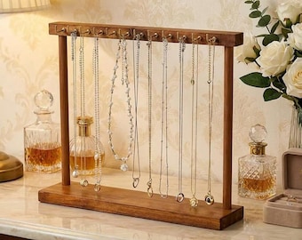 May include: A rectangular wooden jewelry stand with a horizontal bar featuring twelve metal hooks. Several gold-toned necklaces, including a beaded chain and a pearl-accented necklace, hang from the hooks. The stand has two vertical supports and a flat rectangular base.