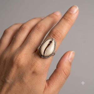 Puede incluir: Un anillo que presenta una concha de cauri de color blanco roto engastada en un marco de plata pulida. El engaste metálico tiene una textura de cuentas en el borde exterior, creando un marco decorativo alrededor de la concha ovalada.