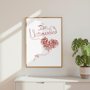 Könnte beinhalten: Ein gerahmter Kunstdruck mit weißem Hintergrund und einer roten Strichzeichnung. Der Text "I'm Unraveled" ist in Schreibschrift gehalten und mit einem Herz aus rotem, verwirrtem Faden verbunden, das unten in einer Nadel endet.