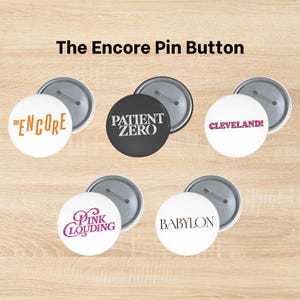 Op de afbeelding: Vijf ronde speldbuttons met metalen achterkant. Eén witte button toont THE ENCORE in oranje, één zwarte button toont PATIENT ZERO in wit, één witte button toont CLEVELAND! in roze, één witte button toont PINK CLOUDING in roze en één witte button toont BABYLON in zwart.