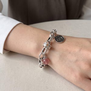 Può includere: Un bracciale a maglie in argento con un ciondolo rotondo raffigurante un quadrante d'orologio, una perla cilindrica incisa con le parole LOVE e RAGE e una perla sferica in pietra rosa traslucida con un cappuccio decorativo in argento.