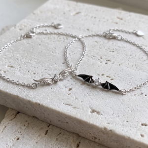 Op de afbeelding: Twee zilverkleurige kettingarmbanden met centrale bedels. De ene armband toont zilveren engelvleugels met een heldere steen, de andere zwarte vleermuisvleugels met een heldere steen. Beide hebben verstelbare kettingen.