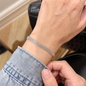 May include: A silver-toned bracelet featuring a fine, woven mesh chain design. The bracelet has a uniform, flexible structure with a metallic finish that wraps around the wrist, showcasing a detailed, interconnected link pattern throughout.