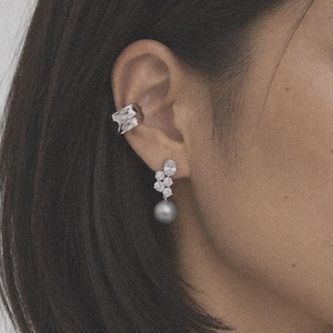 Op de afbeelding: Een zilverkleurige ear cuff met een textuurrijk, onregelmatig oppervlak en een hangende oorbel met een cluster van drie kleine, heldere, rondgeslepen stenen boven een enkele, gladde, ronde, grijze parel die aan de oorlel hangt.