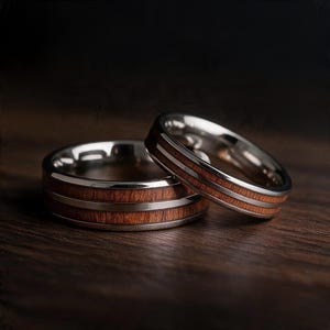 May include: Two rings featuring a polished silver-tone metal band inlaid with a strip of warm brown wood grain. The rings have a smooth, reflective finish and a distinct layered design where the wood is centered between two metal edges.