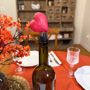 May include: A wine bottle stopper featuring a hand-carved, textured, bright pink heart topper attached to a silver-toned metal base with black rubber rings, inserted into a dark glass Cakebread Cellars wine bottle with a white bear graphic.