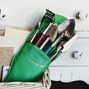May include: A green plastic holder labeled HOME GARDEN SET containing a collection of metal gardening tools, including a small rake, a trowel, and a spade with wooden and metal handles, tucked into a white woven basket.