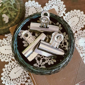 May include: Three silver-toned metal binder clips with the text Boston Clip No. 3 embossed on the surface. The clips are arranged inside a circular, dark green, wire-wrapped container that features a visible coiled structure throughout its frame.
