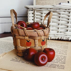 May include: A small woven basket with two handles, featuring painted red apple motifs on the exterior. The basket is filled with several smooth, dark red miniature apple-shaped ornaments, with three additional ornaments resting in front.