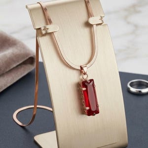 May include: A rose gold-toned herringbone chain necklace featuring a rectangular pendant with a deep red faceted gemstone. The pendant is encased in a decorative metal frame with intricate side detailing, suspended from a simple metal bail.