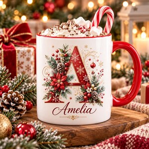 May include: A white ceramic mug with a red handle featuring a large red letter A decorated with poinsettias, pine branches, pinecones, and berries. The name Amelia is printed in red script below the letter. The mug is filled with whipped cream and a candy cane.