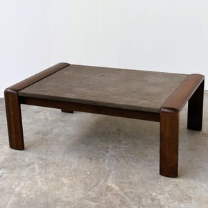 May include: A rectangular coffee table featuring a dark brown wooden frame with rounded edges and a flat, textured, grey stone-like tabletop surface. The structure consists of four sturdy legs connected by horizontal wooden supports.