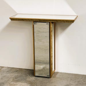 Könnte beinhalten: Ein Konsolentisch mit einer rechteckigen weißen Tischplatte und einer goldfarbenen, strukturierten Kante. Der Sockel ist eine vertikale, rechteckige Säule mit Spiegelflächen an der Vorderseite und den Seiten, eingefasst in goldfarbene Leisten.