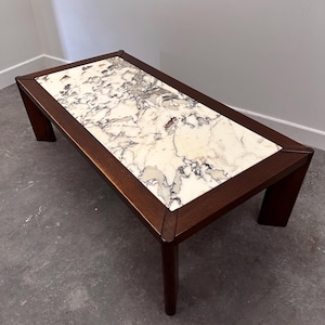 Op de afbeelding: Een rechthoekige salontafel met een donkerbruin houten frame met vier stevige poten en een glad, ingelegd tafelblad van wit marmer met opvallende grijze en zwarte aders die door het gehele steenmateriaal lopen.