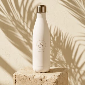 Könnte beinhalten: Eine cremeweiße Trinkflasche mit mattem Finish und einem silberfarbenen Metalldeckel. Die Flasche ist mit einem kreisförmigen Emblem versehen, das den Buchstaben S und den Namen SOPHIE in einer dünnen, dunklen Schrift mittig zeigt.