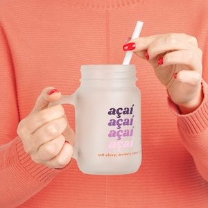 Könnte beinhalten: Ein Einmachglas-Becher aus Milchglas mit Henkel und weißem Strohhalm. Auf der Vorderseite ist das Wort "açaí" viermal in einem vertikalen Farbverlauf von Dunkellila bis Hellrosa aufgedruckt, gefolgt vom Text "all day, every day" in Orange.