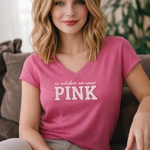 Op de afbeelding: Een T-shirt met korte mouwen en een V-hals in een felroze kleur. De voorkant van het shirt is voorzien van de witte tekst "in october we wear" boven het woord "PINK" in een groot bloklettertype met een wit en roze ruitpatroon.