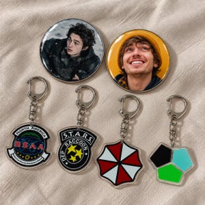 Op de afbeelding: Twee ronde buttons met portretten van personen en vier acryl sleutelhangers. De sleutelhangers tonen het BSAA North America logo, het S.T.A.R.S. Raccoon Department embleem, een rood-wit paraplusymbool en een geometrische vorm.