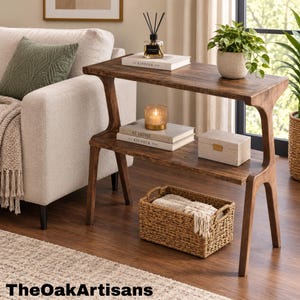 May include: A two-tier wooden console table with a dark brown finish, featuring a top shelf holding a reed diffuser and a potted plant, and a lower shelf with two stacked books, a small rectangular box, and a woven basket underneath.