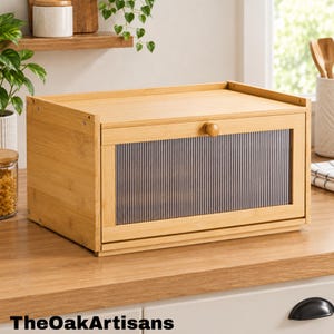 May include: A rectangular bamboo bread box featuring a drop-down door with a fluted, semi-transparent acrylic panel and a small round wooden knob. The natural light-toned wood grain is visible across the exterior frame and top surface.