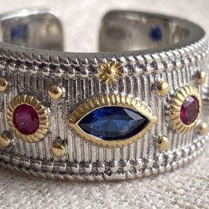 May include: A wide silver-toned open band ring featuring a textured, vertically striped surface. The ring is adorned with a central marquise-cut blue gemstone, two round red gemstones, small gold-toned star accents, and raised gold-toned dots.