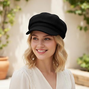 May include: A solid black newsboy cap featuring a structured crown, a short stiff brim, and a band detail across the front. The hat is crafted from a textured fabric and maintains a rounded shape that sits comfortably on the head.