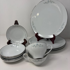 May include: A white porcelain dinnerware set featuring a delicate pattern of small blue flowers and green leaves with silver rims. The collection includes dinner plates, bowls, and teacups, all displaying a consistent floral motif.