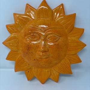 May include: A circular ceramic wall plaque shaped like a sun with a central face featuring almond-shaped eyes, a straight nose, and closed lips. The surface is glazed in a vibrant orange hue with dark brown speckles throughout the rays and face.