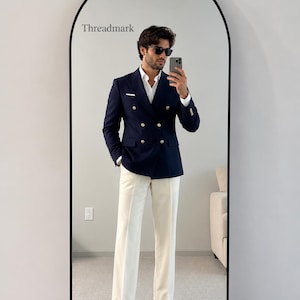 Könnte beinhalten: Ein marineblaues zweireihiges Sakko mit goldenen Knöpfen, ein weißes Hemd mit Kragen und eine cremefarbene Hose. Das Outfit wird durch dunkelbraune Leder-Loafer und eine schwarze Sonnenbrille ergänzt. Das Wort Threadmark ist auf dem Spiegel zu sehen.