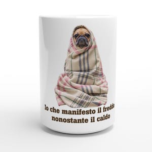 Op de afbeelding: Een witte keramische mok met een afbeelding van een beige mopshond gewikkeld in een beige, roze en zwart geruite deken. Onder de hond staat de tekst: Io che manifesto il freddo nonostante il caldo. De mok heeft een cilindrische vorm.