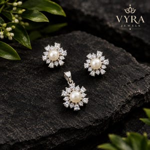 Peut inclure: Un ensemble de bijoux composé de deux boucles d'oreilles et d'un pendentif assorti. Chaque pièce présente une perle blanche ronde centrale entourée d'un halo de pierres claires en forme de poire sur métal argenté. Logo Vyra Jewels visible.