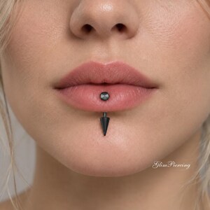 May include: A black lip piercing jewelry piece featuring a flat circular top disc resting on the upper lip and a pointed cone-shaped spike extending downward from the lower lip. The metal has a matte black finish and the text Glim Piercing.