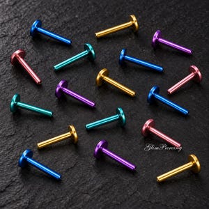 Puede incluir: Una colección de piercings labret de base plana en varios colores metálicos, incluyendo azul, dorado, morado, turquesa y rosa. Cada pieza presenta un disco circular liso y un poste recto. El texto GlimPiercing es visible en la esquina inferior.